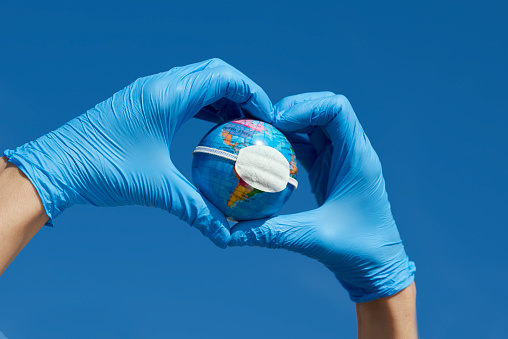 man, wearing blue surgical gloves, forming a heart with his hands, holding a terrestrial globe with a protective mask, depicting the global coronavirus pandemic or the air pollution in the planet